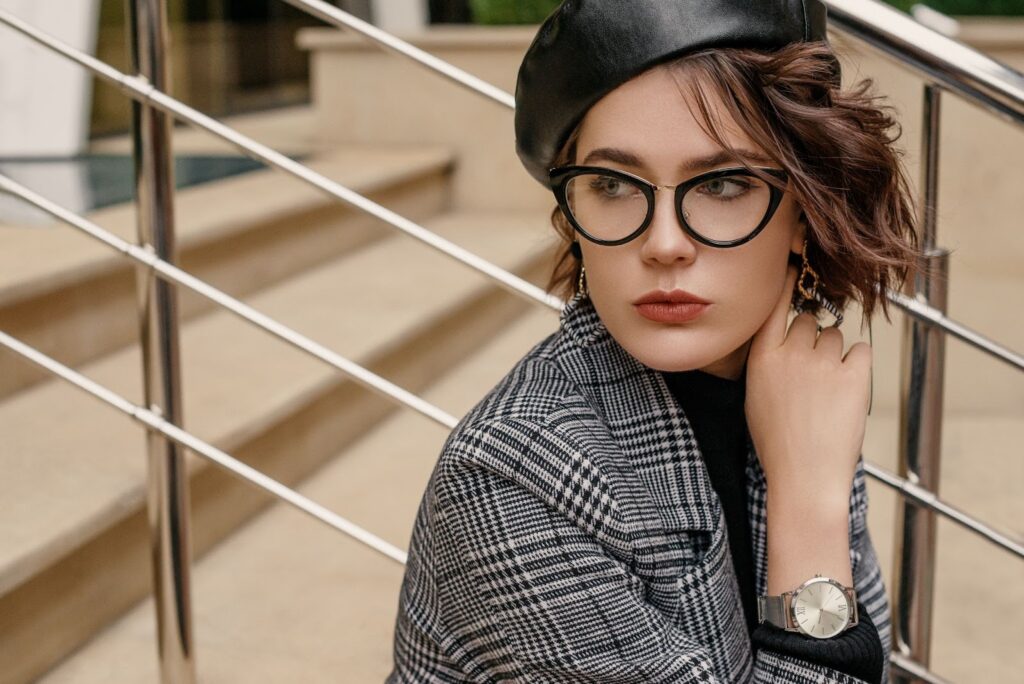Stylish woman posing with black cat-eye eyeglass frames, black asymmetrical hat, silver watch, and black and white plaid jacket.